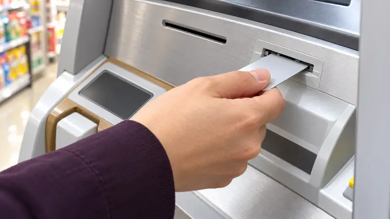 Illustrative close-up of a traveller inserting a bank card into an ATM inside a Japanese convenience store.