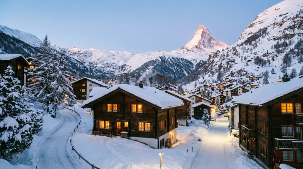 Snow-covered traditional wooden chalets in a Swiss Alpine village at dusk during winter.