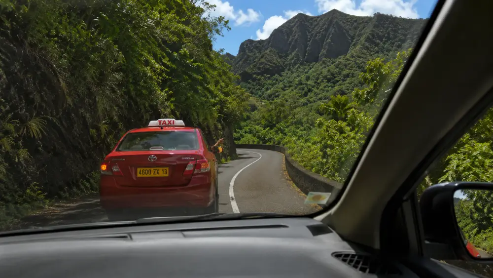 View from a car passenger seat on a narrow, winding road in Mauritius, showing an official taxi driving ahead through tropical mountains.