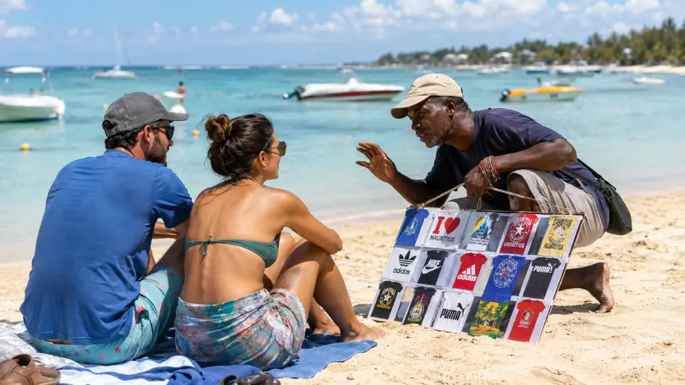 A persistent beach vendor attempts to sell souvenirs and fake branded clothing to a tourist couple on a public beach in Mauritius.