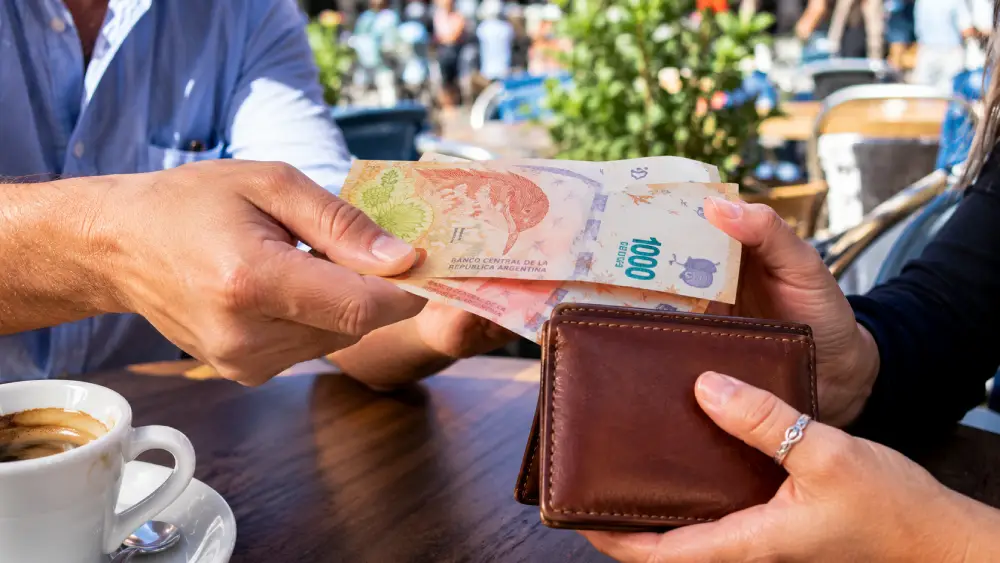 Close up of hands exchanging bright Argentine currency at a sunny outdoor cafe.