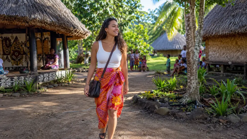 A solo female traveler wearing a traditional colorful sulu walking confidently through a sunny, welcoming village in Fiji.
