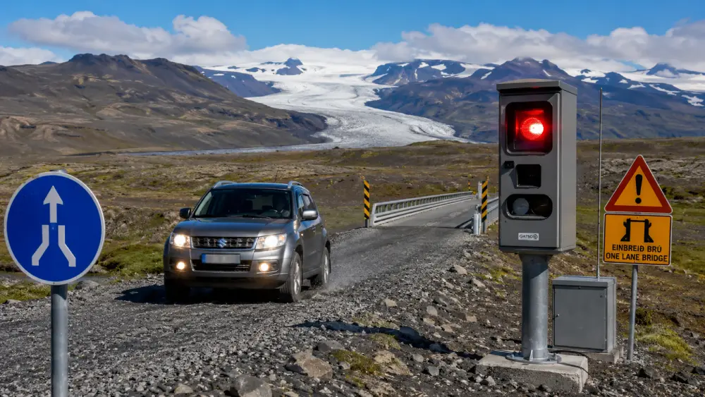 A rental 4x4 drives past a flashing speed camera on a winding Icelandic mountain road, with a single-lane bridge merging sign visible in the foreground.