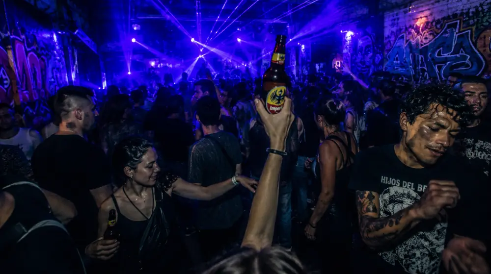 A raw, atmospheric photograph capturing the kinetic energy of an underground electronic music club in San Jos&eacute;'s La California neighborhood, featuring a dancing crowd bathed in deep blue and purple neon lights, graffiti-covered brick walls, and a thick haze of smoke.