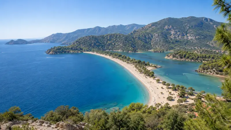 Turquoise water and the curved beach of Ölüdeniz’s Blue Lagoon near Fethiye, surrounded by green hills.