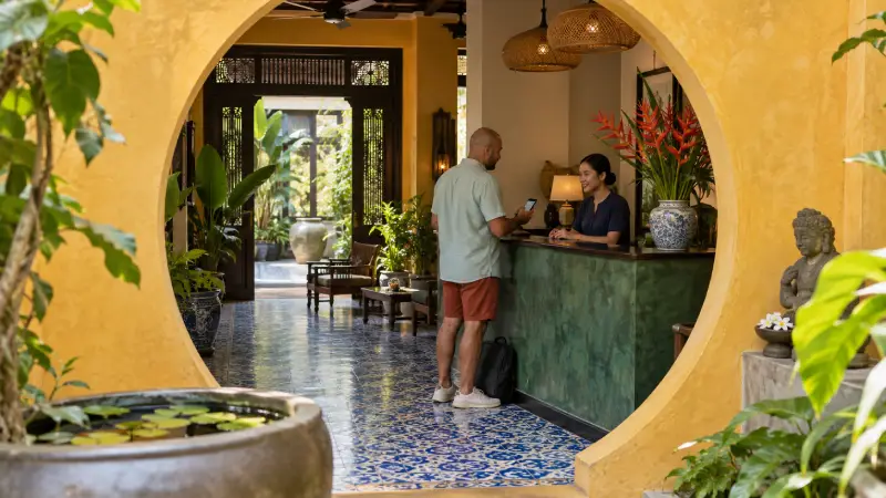 Pengunjung memeriksa reservasi hotel di meja resepsionis hijau yang dibingkai bukaan halaman berbentuk lingkaran.