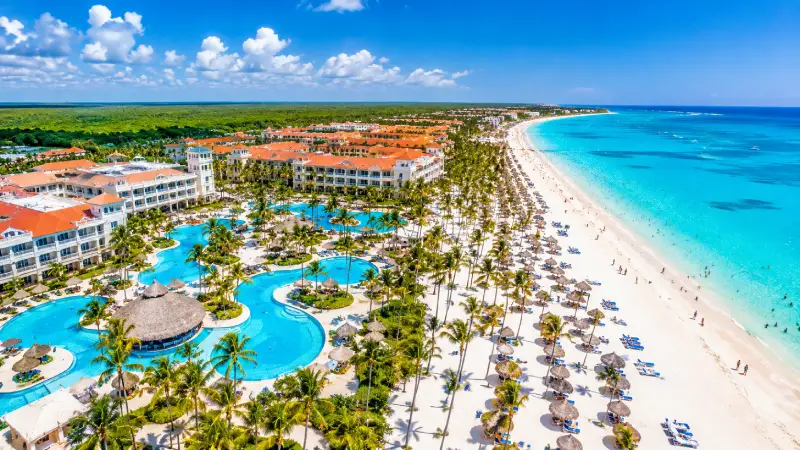 Crowded white sand beach and luxury all-inclusive resorts in Punta Cana lined with palm trees under bright sun.