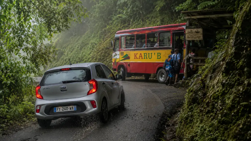 Une voiture de location argentée navigue dans un virage abrupt et brumeux de la forêt tropicale tandis qu'un bus public coloré Karu'B prend des passagers à un abri rustique en Guadeloupe.