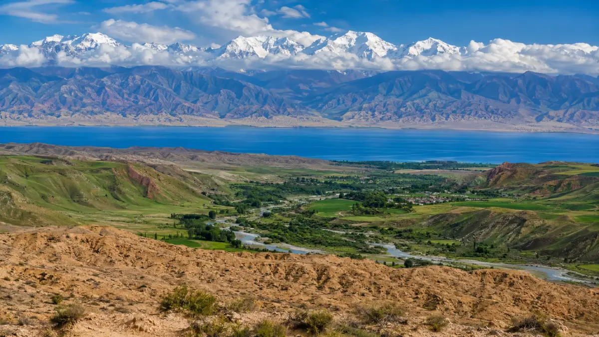 A wide landscape photo showing diverse Kyrgyz geography simultaneously: arid southern plains, lush central valleys, the large Lake Issyk-Kul, and high, snow-capped Tian Shan mountains.