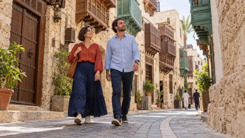Adult travelers in loose, modest sightseeing clothes walking through an illustrative historic Jeddah lane.