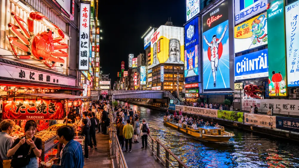 A dynamic nighttime shot of the Dotonbori Canal in Osaka, famous for its colorful neon signs, including the Glico Running Man and lively street food culture.
